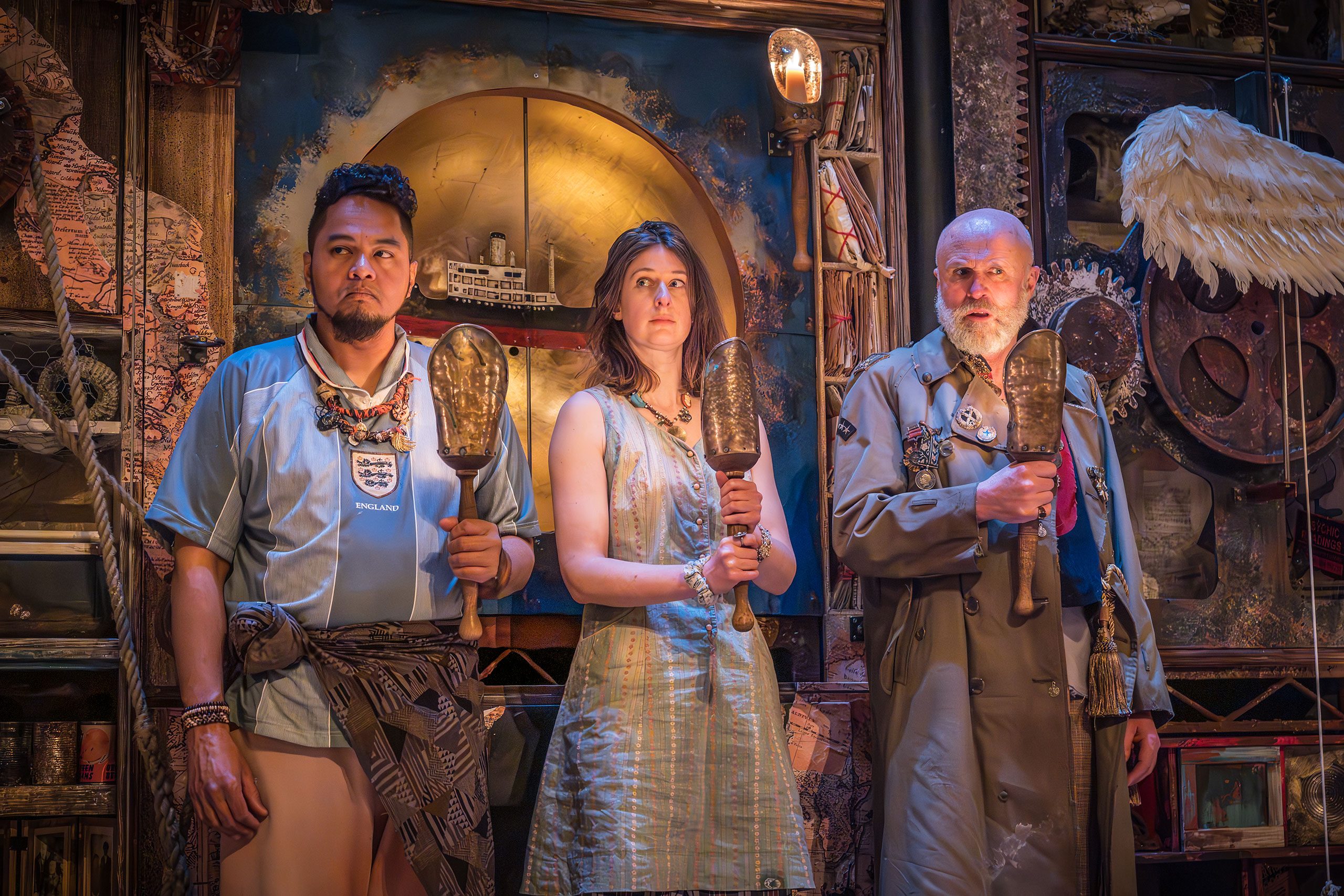 Three people stand side by side, each holding golden candlesticks. One of them wears a light blue football top with an England badge and beads around their neck. The other wears a pale striped dress. The third person wears a grey trench coat with military badges and tassels on it. The wall behind them is covered in various objects: books, gears, an angel wing, ropes and a model of a boat.