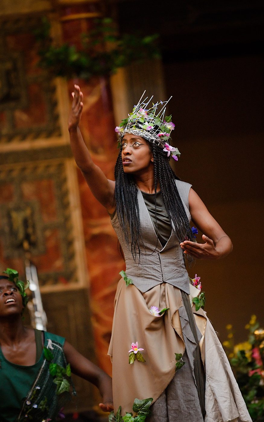An actor performs on stage wearing a crown of flowers, with one arm lifted in the air