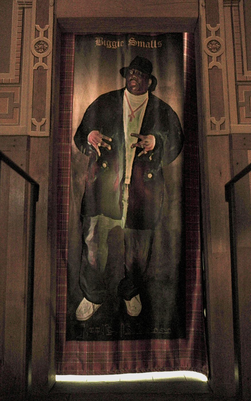A tapestry of a person standing, wearing a black hat, white polo neck, black jacket and trousers and white trainers with a gold cross around their neck. The tapestry has patterned edges and hangs within a wooden doorframe.