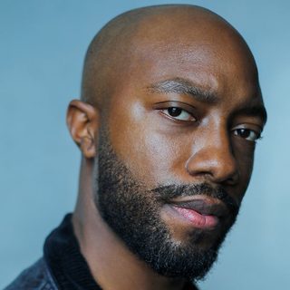 Elijah Cook looking directly at the camera. They are wearing dark clothing in front of a blue background.