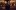 An actor wearing a crown facing away from the camera towards a banquet table on stage with blurred outlines of other actors at the table