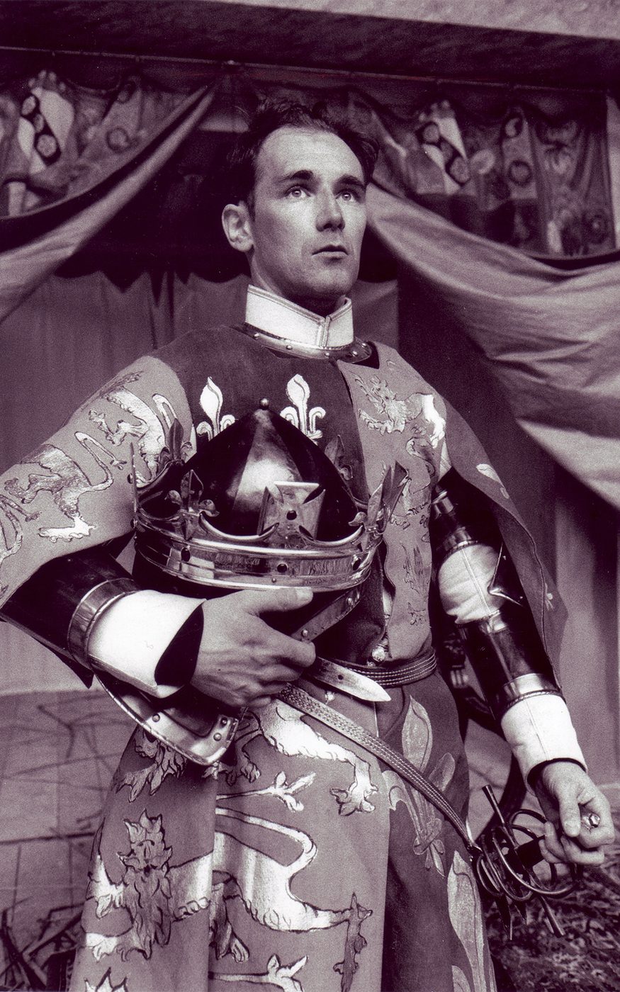 A man wearing a tunic with the English and French emblems holds his crown under his arm.