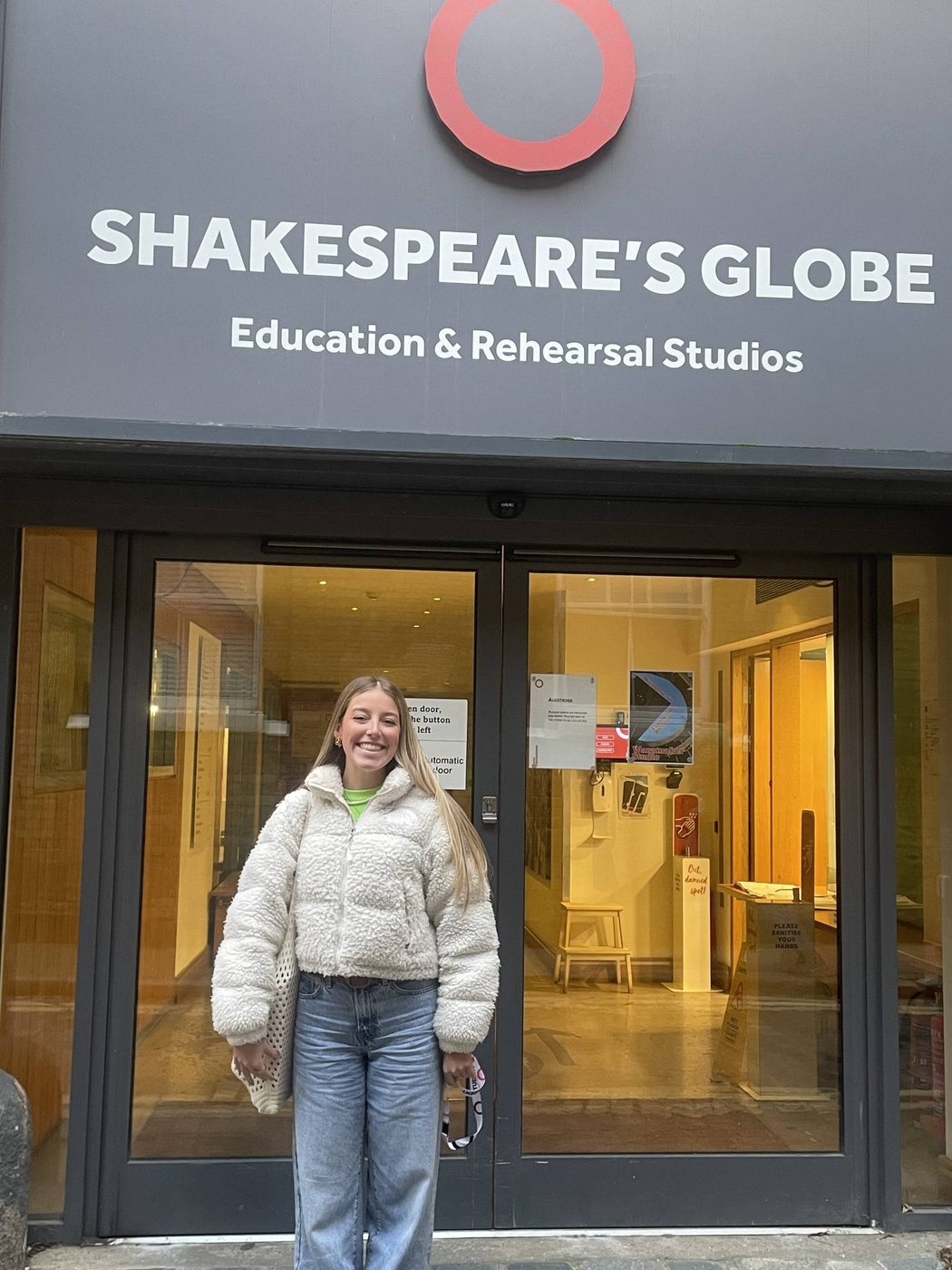 CJ, our MA Shakespeare Studies Bursary student, standing and smiling outside of Shakespeare's Globe Education & Rehearsal Studios. They have long blonde hair and are wearing a white puffer jacket and blue jeans.