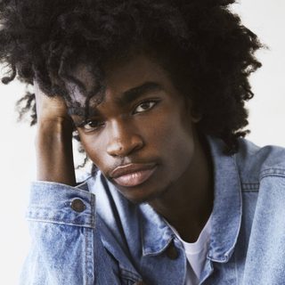 Jayden Elijah looking directly at the camera. They are wearing a blue denim jacket and leaning their head against their arm.
