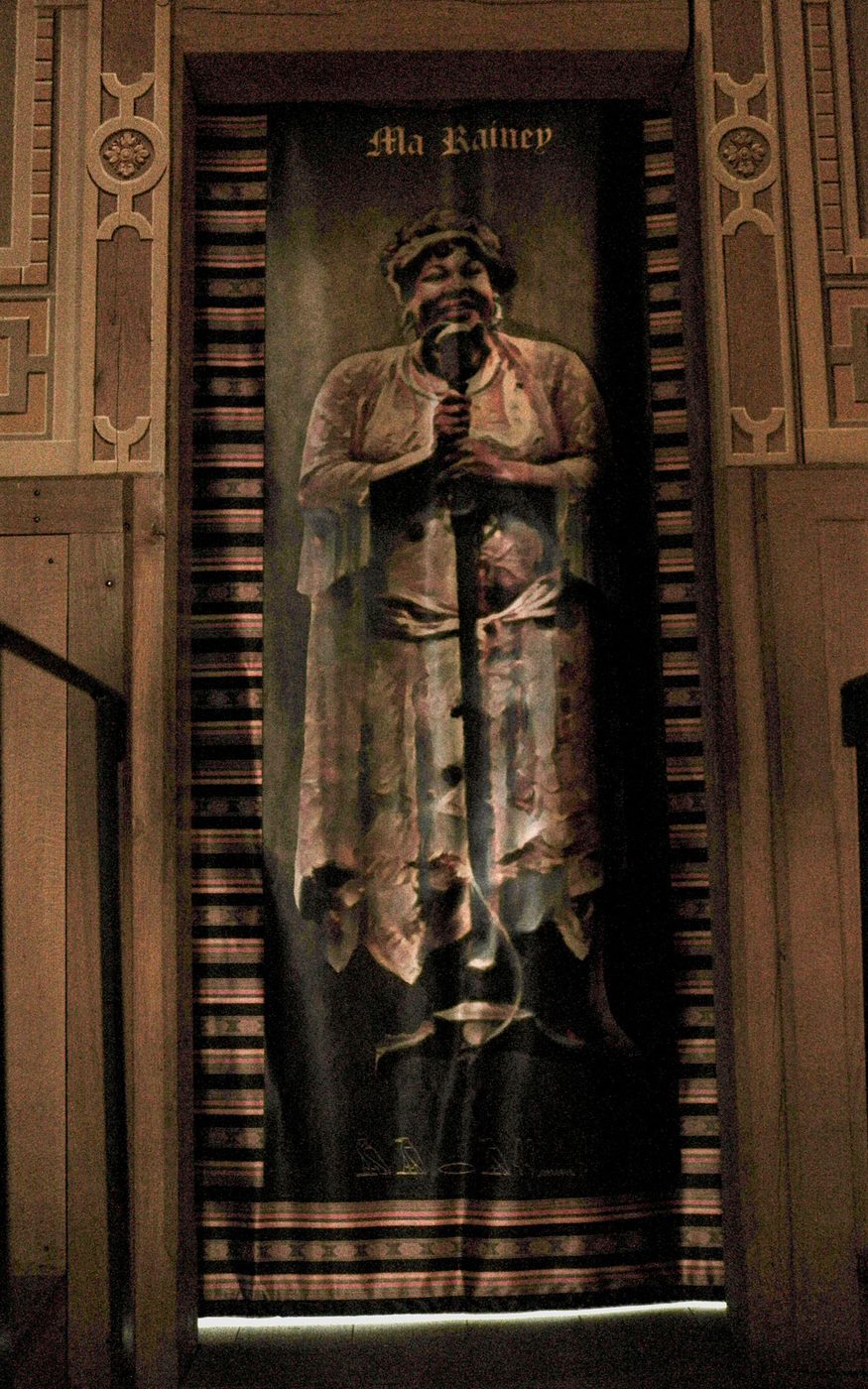 A tapestry of a person wearing a long cream dress and cap, standing behind a microphone. The tapestry has patterned edges and hangs within a wooden doorframe.