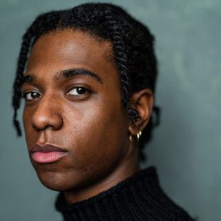 Maxwell Chartey looking directly at the camera. He has short black hair and is wearing a black turtle neck jumper.