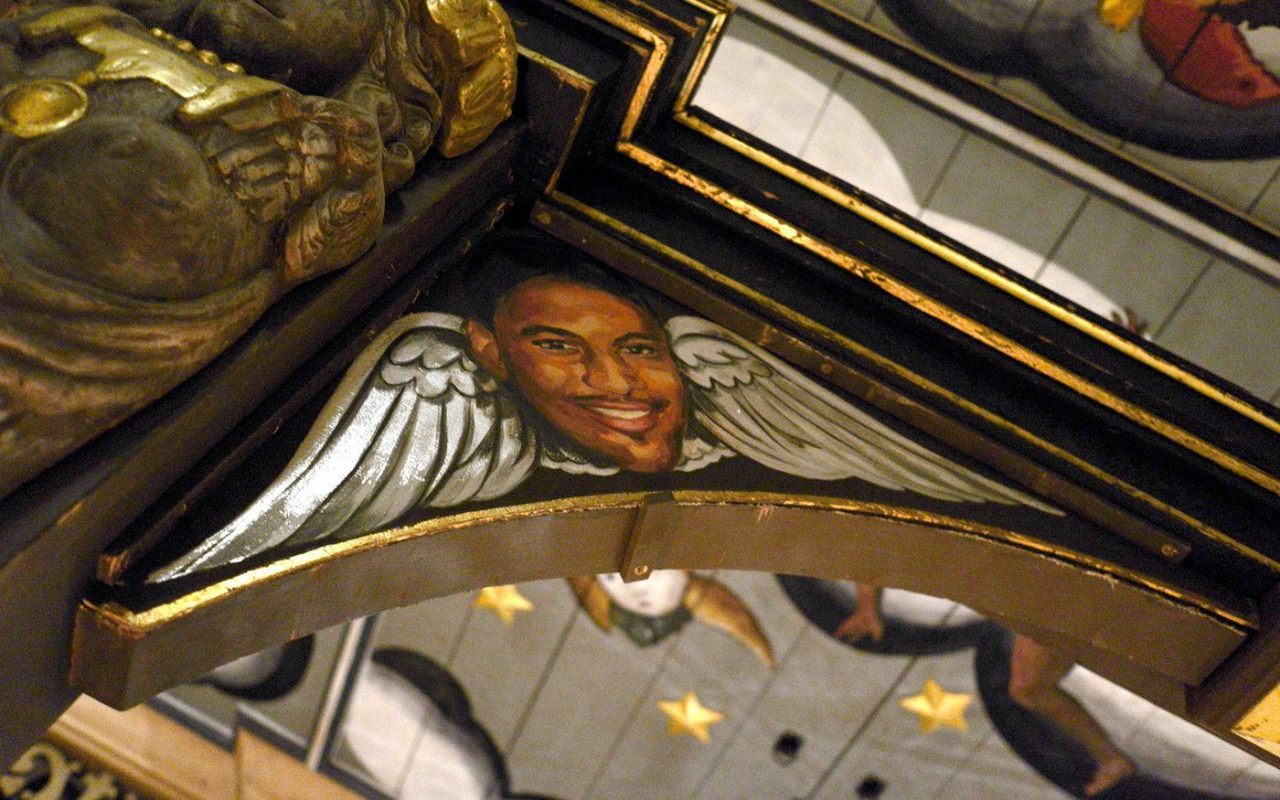 A face with angel wings on either side of it, painted on a triangular wooden panel of a ceiling with gold edges.