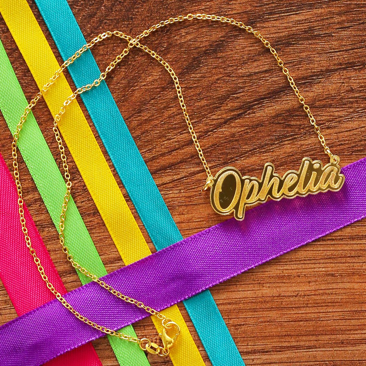 A gold necklace with a mirror pendant reading 'Ophelia' is framed by colourful ribbons.