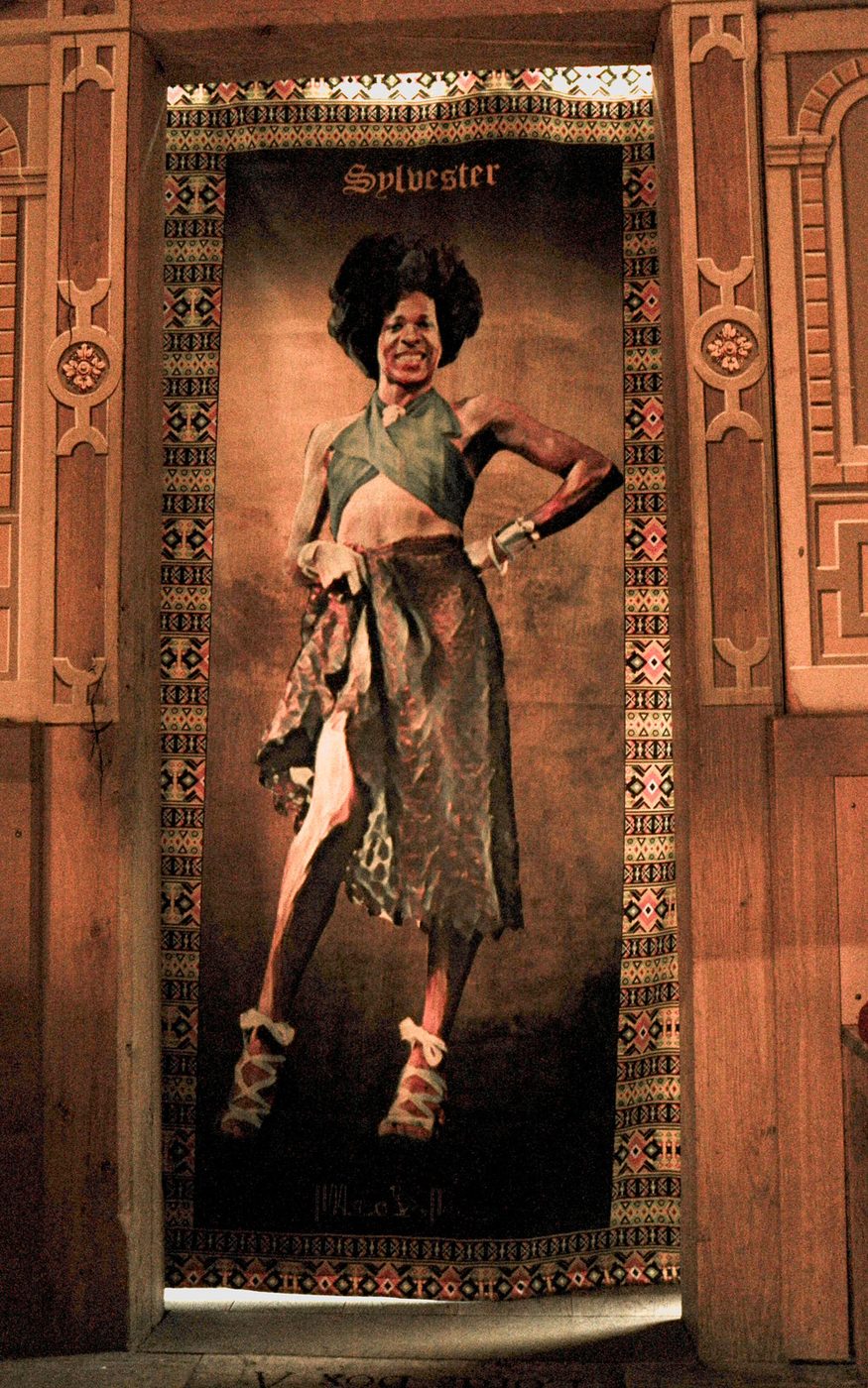 A tapestry of a person standing, smiling, with their hand on their hip, wearing a green cropped top, a patterned skirt and white heeled sandals. The tapestry has patterned edges and hangs within a wooden doorframe.