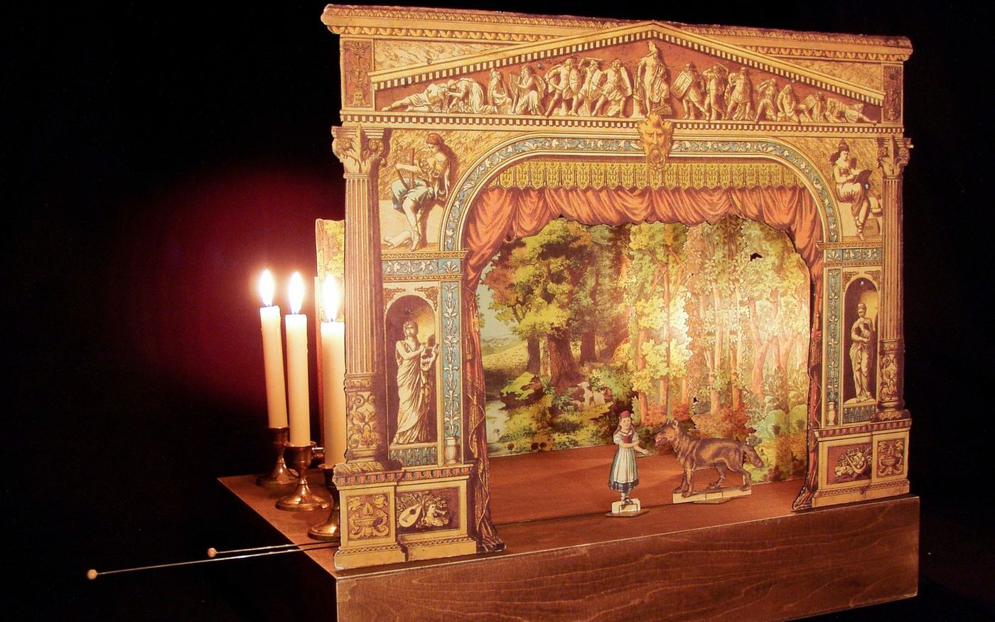 A 3D model of a theatre, made out of paper. The front of the model is an elaborate archway with columns and figures sketched onto it. Behind this are paper scenery of trees and a lake. On the paper stage, there is a paper girl and a paper wolf.