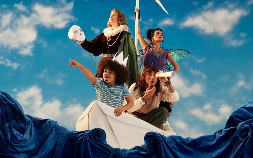 Four people are aboard a giant paper boat against a bright blue, cloudy sky and a sea of dark blue material. Two are standing; one in Shakespearean dress holding a paper skull, another in a purple fairy costume holding a paper mast. The other two are crouched, with one wearing a paper hat, pointing out to sea, and another looking through a paper telescope, scratching their chin.
