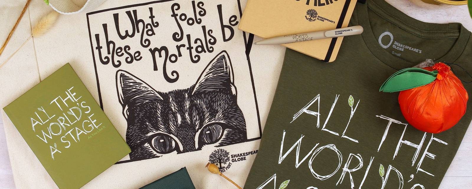 Shakespeare merch including a tote bag, notebooks, t-shirt, pen, and plant holder are displayed on a white wood background surrounded by plants.
