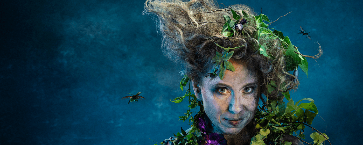 A close up of a smirking woman against a blue backdrop. She is covered in dirt and foliage and has insects flying around her unruly hair.