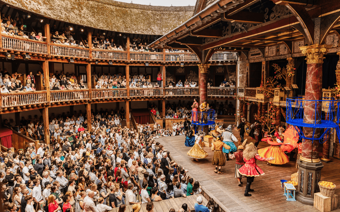 A photograph of the Globe Theatre during a performance. On stage are a group of actors dancing in colourful costumes. The auditorium is full of people standing and sitting.